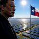 Elon Musk stands in profile against a clear blue sky, with a bright overhead sun shining above a vast landscape of Tesla solar panels stretching to the horizon, and a smaller Texas flag flying on a pole to his right.
